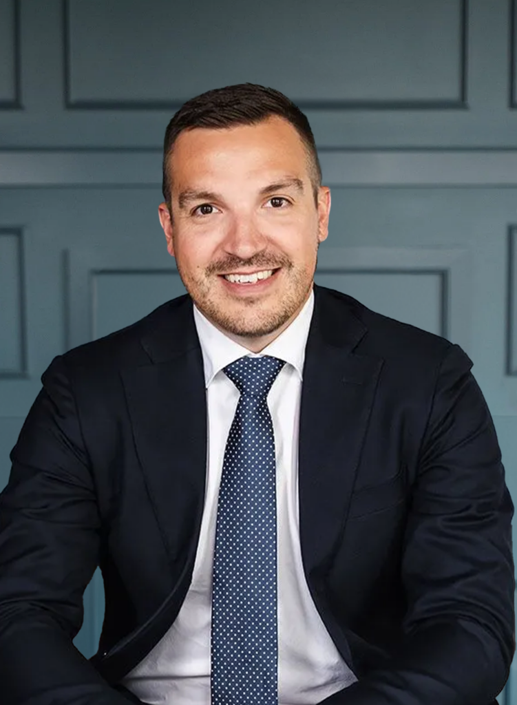 Smiling man in a suit with a dotted tie, sitting against a paneled blue background.