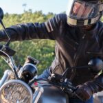 Motorcyclist in leather jacket and helmet riding outdoors, surrounded by greenery.