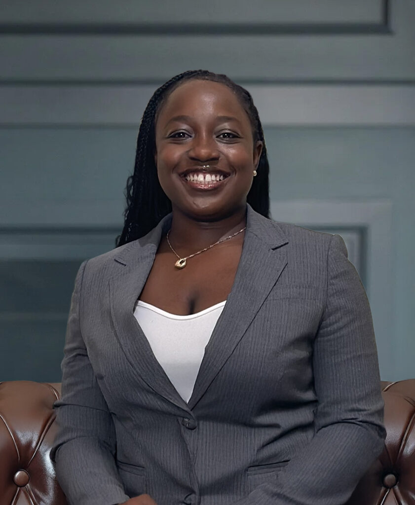 Smiling woman in a gray suit jacket sitting on a brown leather couch in a professional setting.