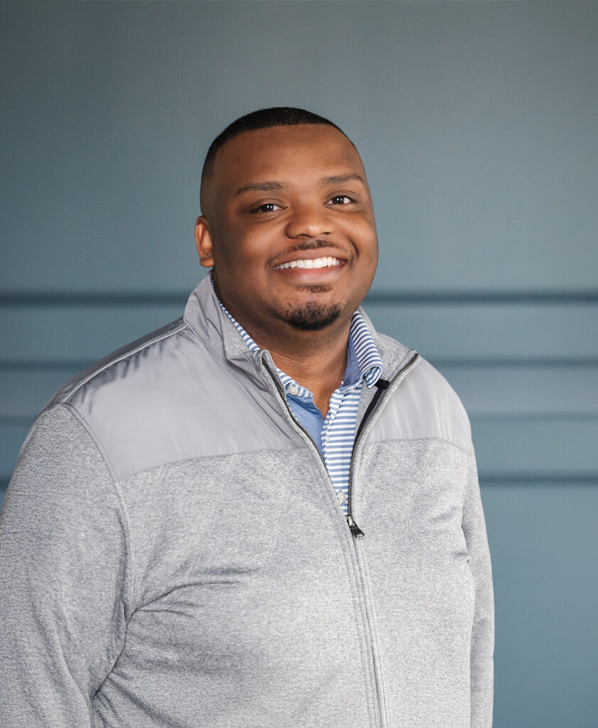 Smiling man in a gray jacket and striped shirt standing against a blue background.