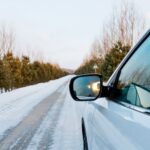 Car driving on a snowy road lined with trees, reflecting the winter landscape in its windows.