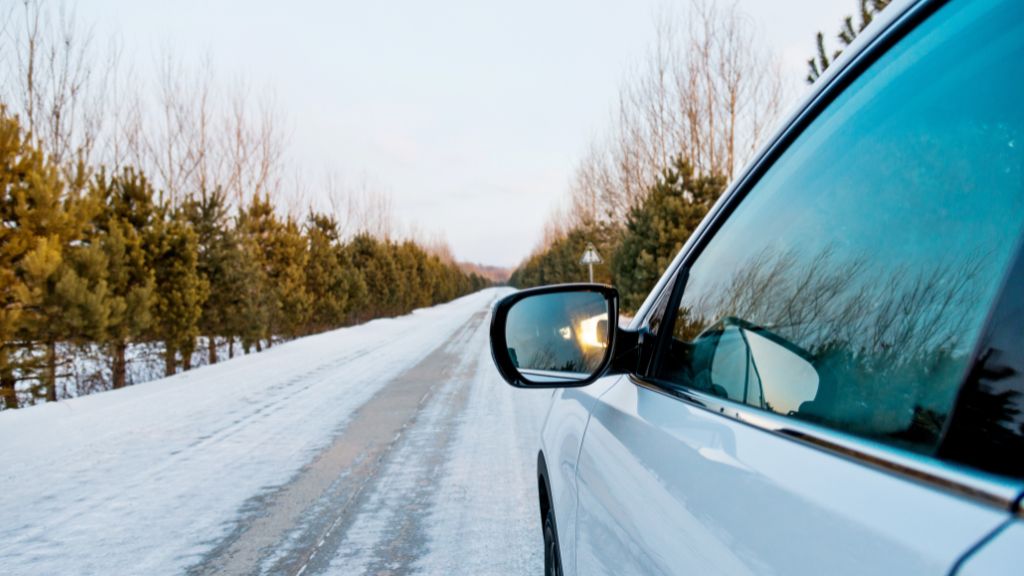 Car driving on a snowy road lined with trees, reflecting the winter landscape in its windows.