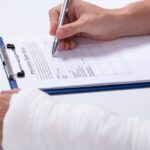 Person with bandaged arm filling out an insurance claim form on a clipboard.