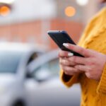 Person in a yellow sweater using a smartphone outdoors near a parked car.