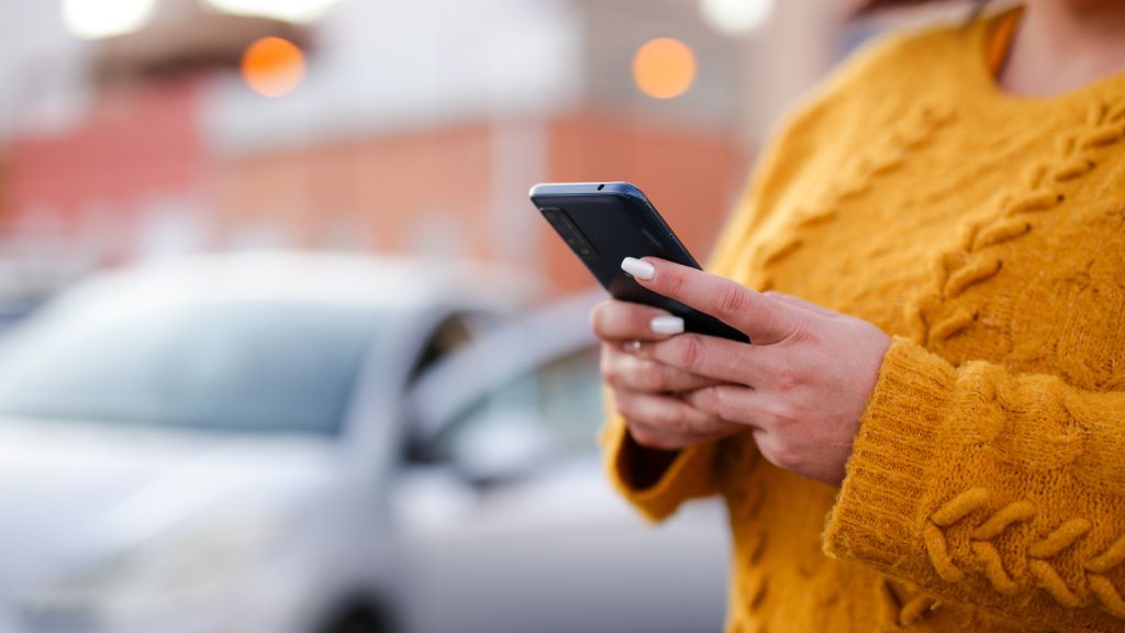 Person in a yellow sweater using a smartphone outdoors near a parked car.
