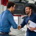 Two men in a car workshop shaking hands, one holding a clipboard, with vehicles in the background.