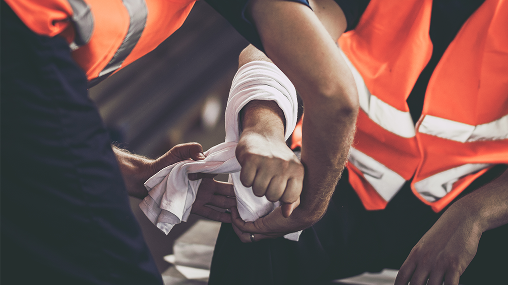Workers in orange safety vests bandaging an injured arm on a construction site.