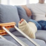 Person with a bandaged leg using a smartphone on a couch; crutches resting nearby.