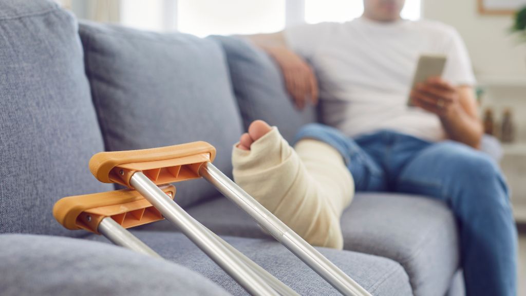 Person with a bandaged leg using a smartphone on a couch; crutches resting nearby.
