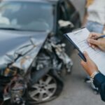 Insurance agent assessing damage on a crashed car with a clipboard, next to an unidentifiable woman in a striped shirt.