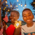 Smiling boy and woman holding sparklers and American flags at a festive outdoor gathering with string lights.