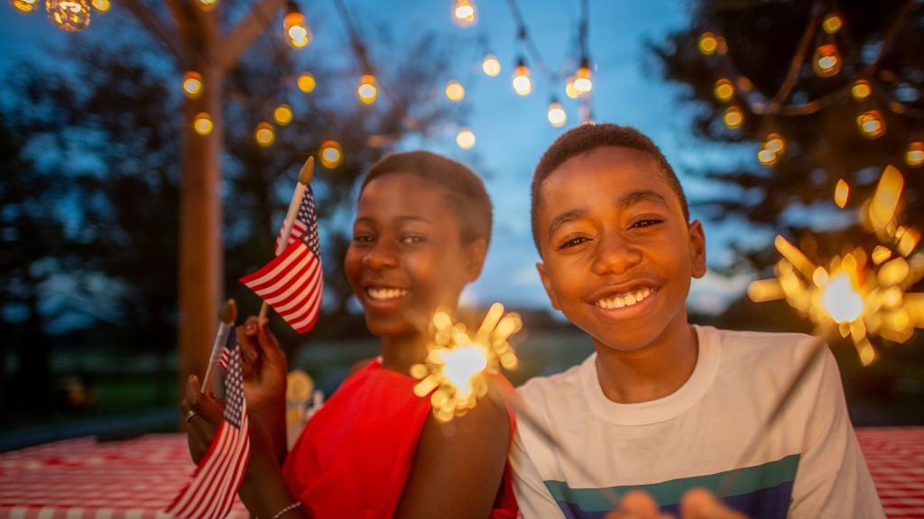 Smiling boy and woman holding sparklers and American flags at a festive outdoor gathering with string lights.