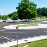Modern roundabout with clear road markings and lush greenery in a suburban area under a bright blue sky.