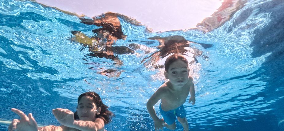 Two children swimming underwater in a pool, surrounded by clear blue water and sunlight streaming from above.