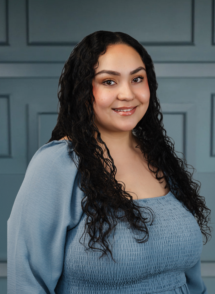 Smiling woman with long, curly hair wearing a blue top, posing against a paneled wall backdrop.