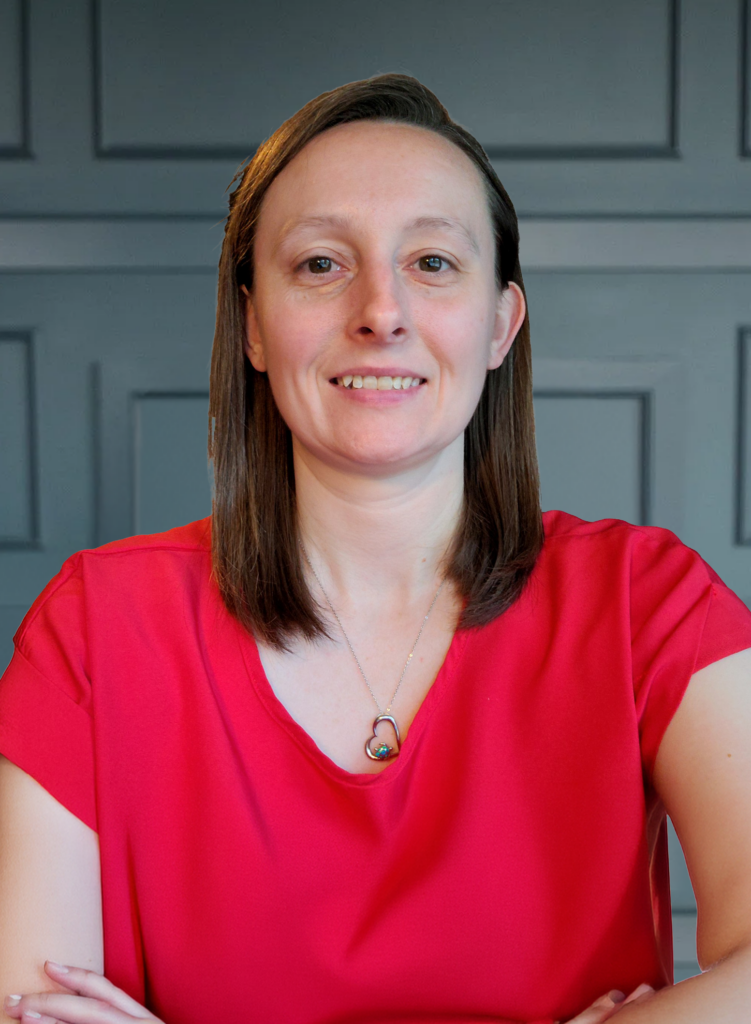 A smiling person in a red shirt with a necklace, posing against a gray paneled background.