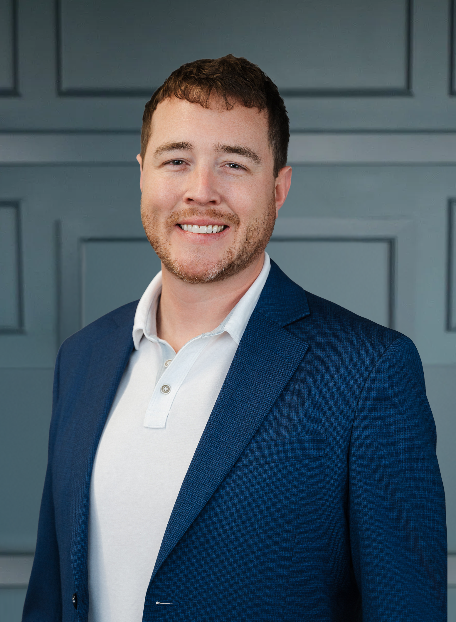 Smiling man in a blue suit and white shirt against a gray paneled backdrop.
