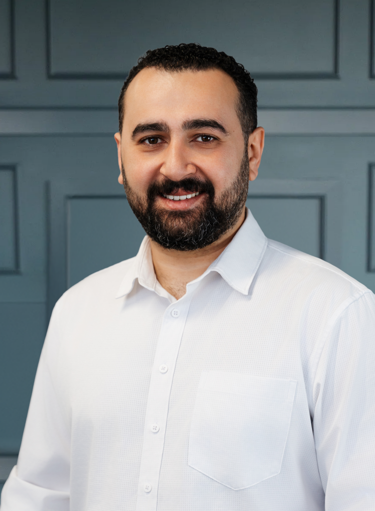 Smiling man in a white shirt with a beard, standing against a blue paneled background.