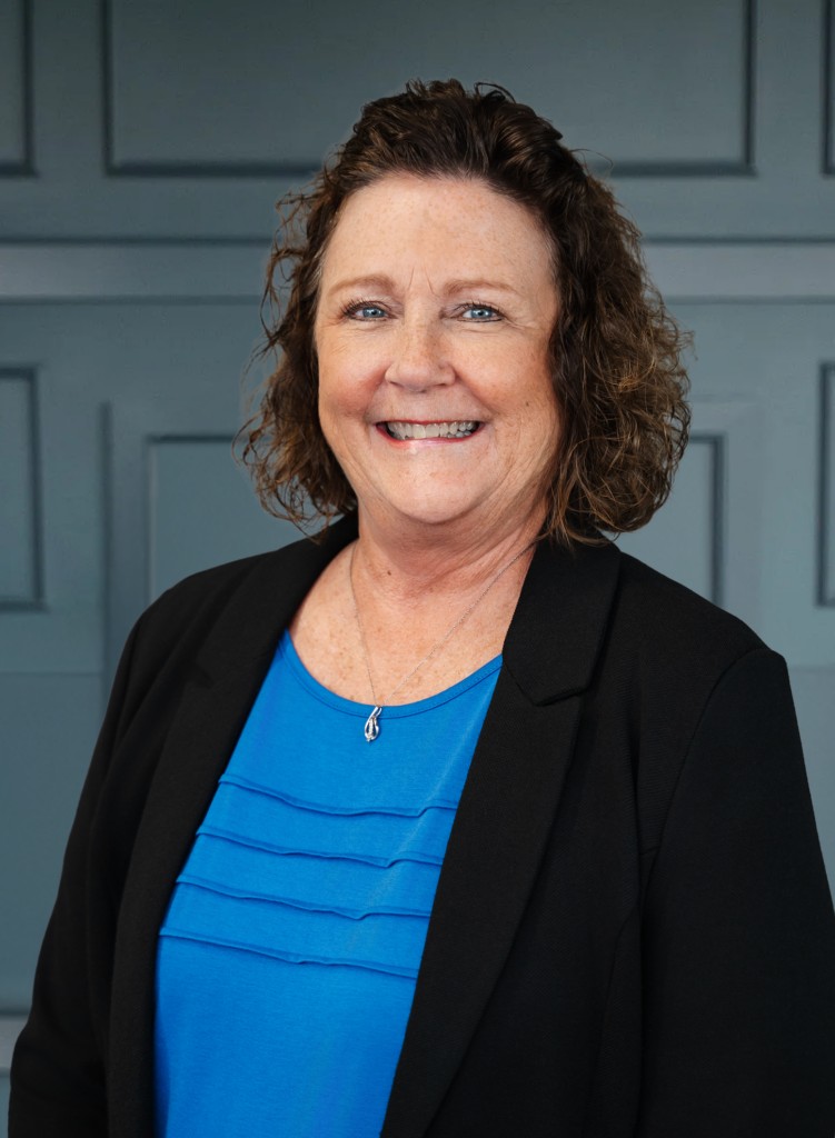 Smiling woman in a blue shirt and black blazer, standing against a paneled wall background.