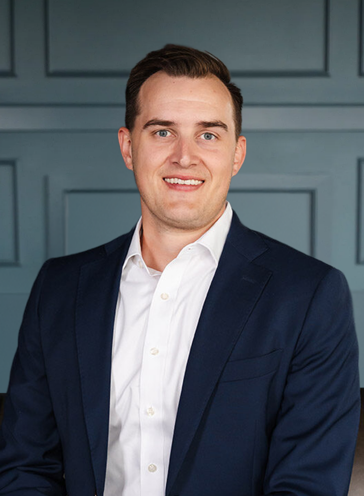 Smiling man in a navy suit and white shirt against a paneled blue background.