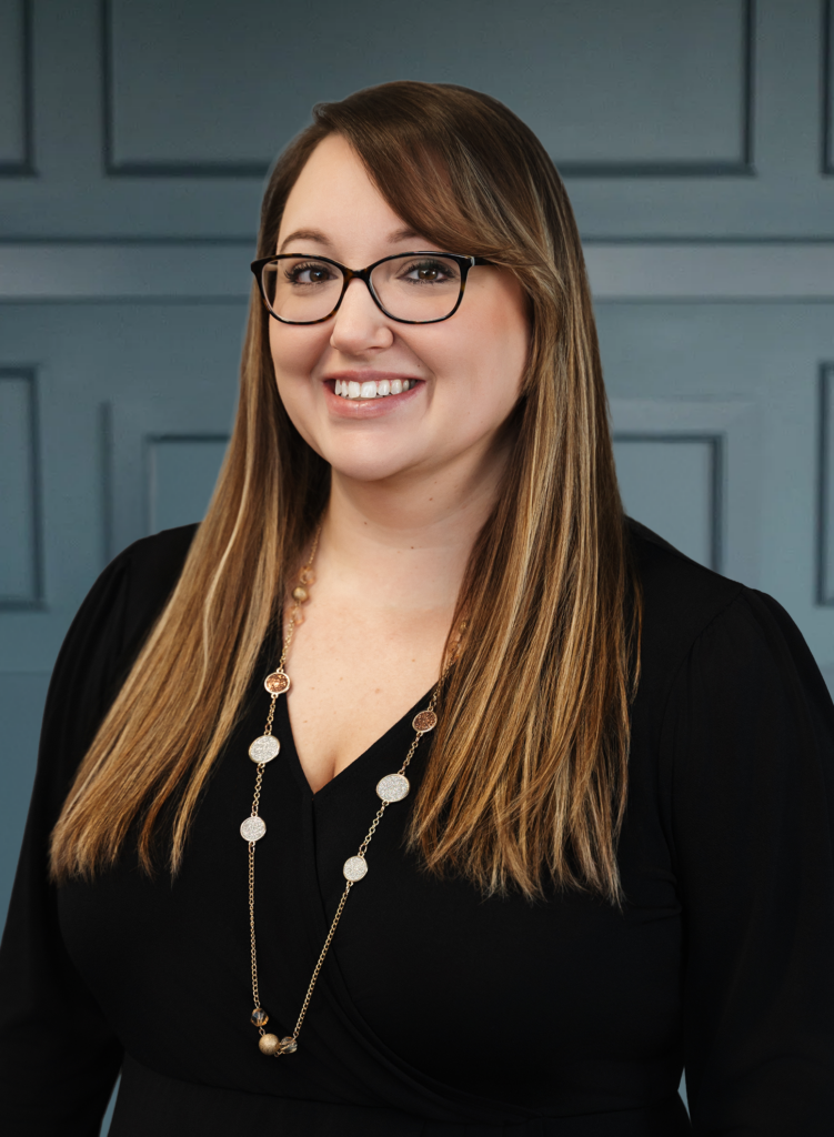 Smiling woman with glasses wearing a black top and necklace, standing against a panelled blue background.