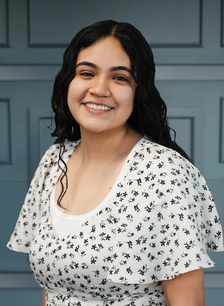 Smiling person with long black hair, wearing a white floral dress, standing in front of a blue paneled background.