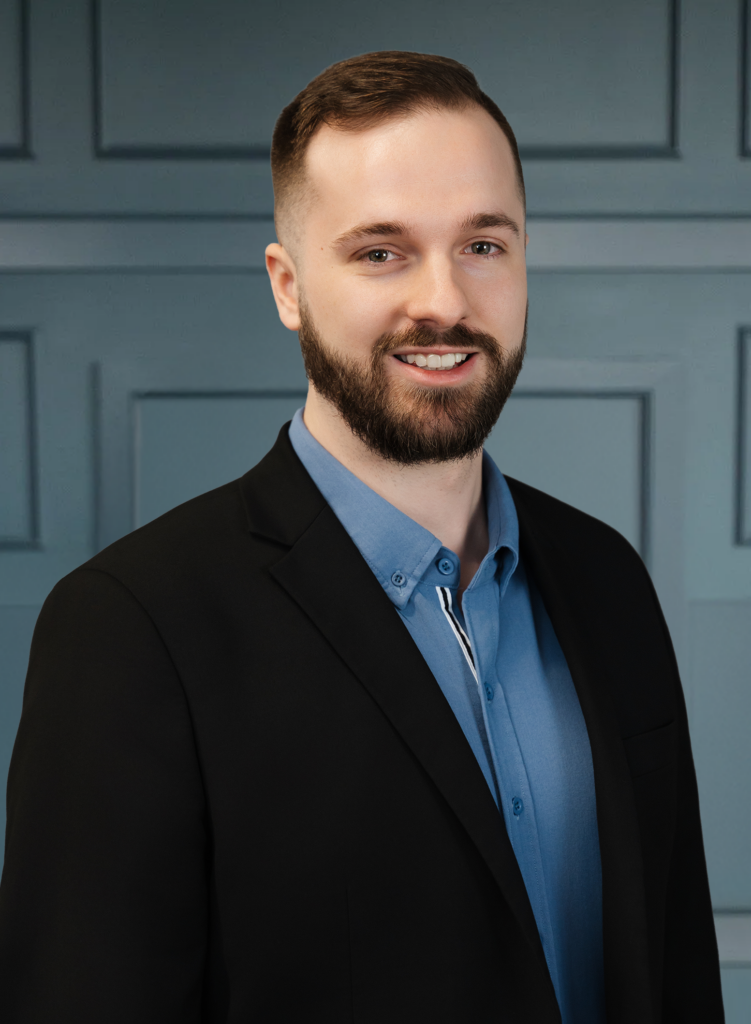 Man in a suit with a beard and short hair, smiling against a blue paneled background.