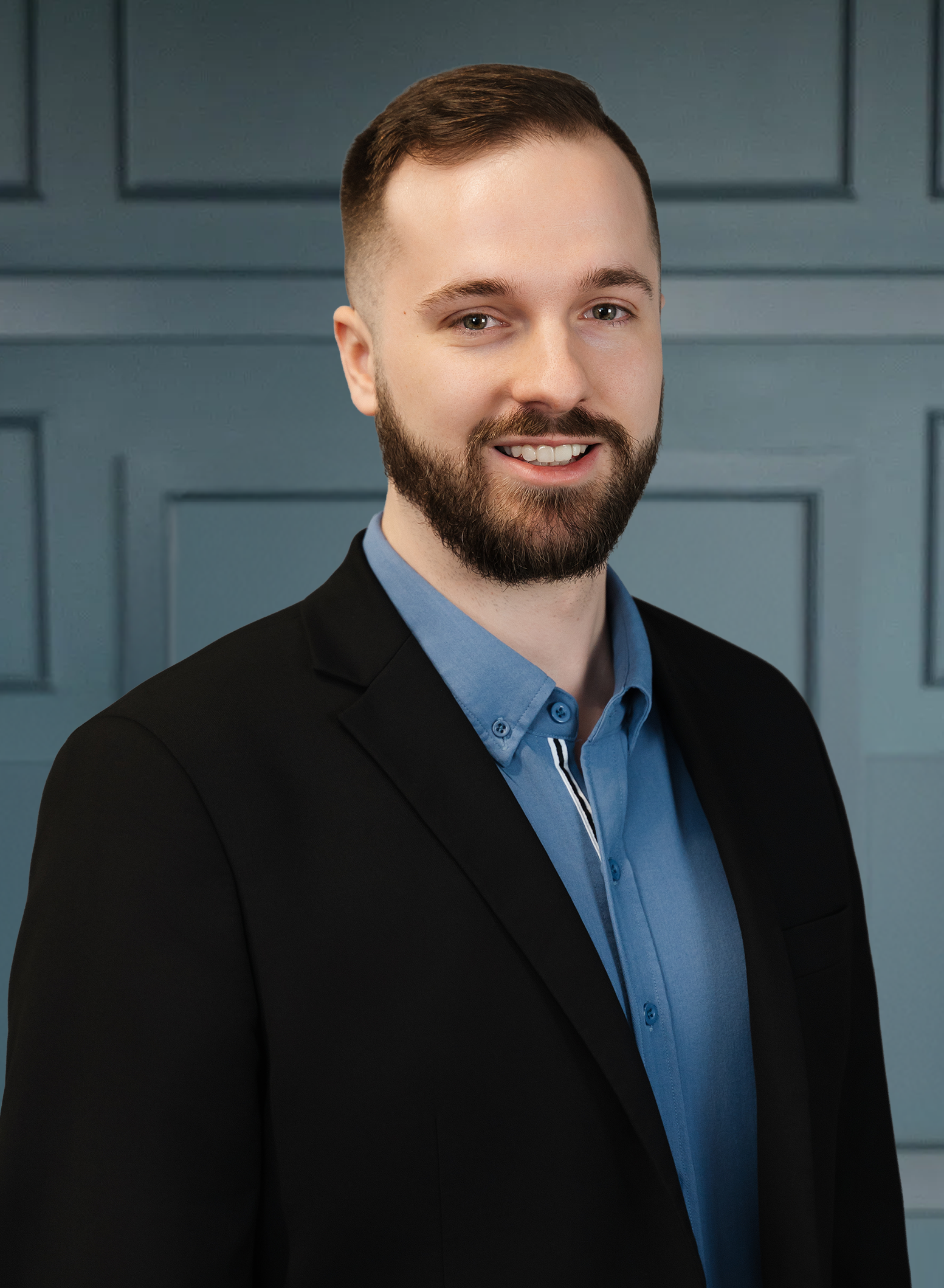 Man in a suit with a beard and short hair, smiling against a blue paneled background.