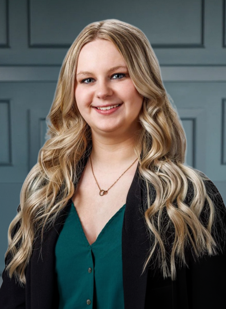 Smiling woman with long blonde hair wearing a green top and black blazer, standing in front of a gray paneled wall.