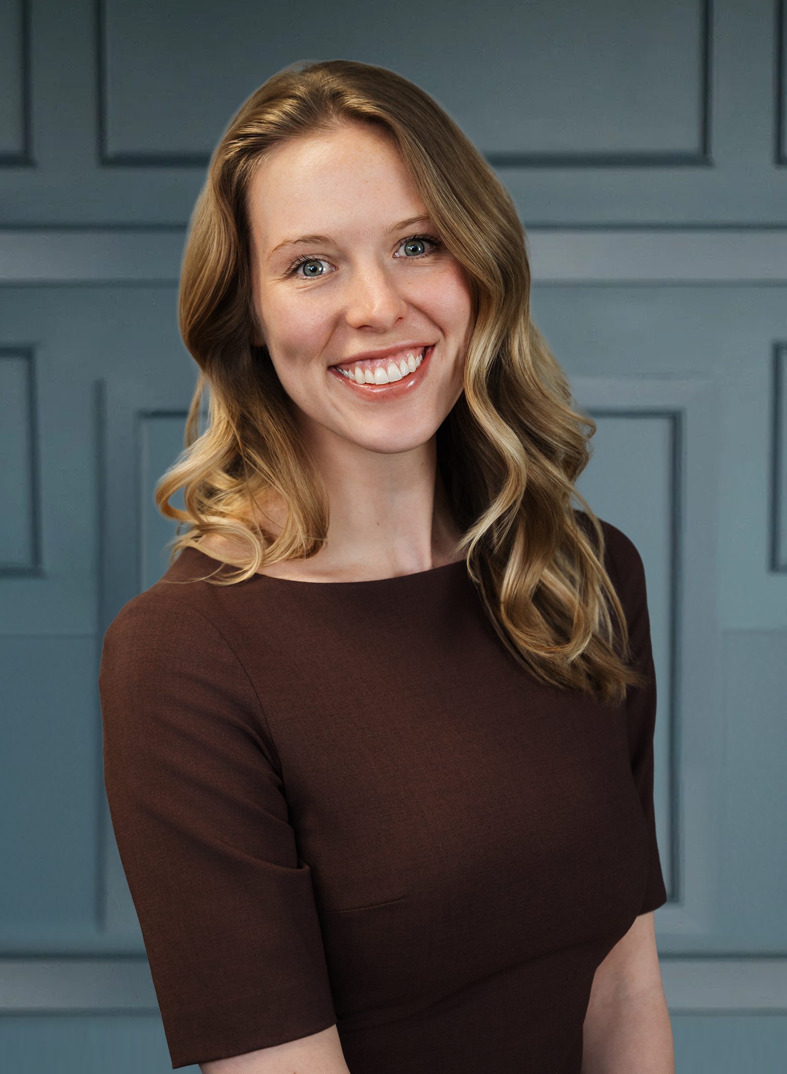 Smiling woman with wavy blonde hair in a brown dress against a blue paneled background.