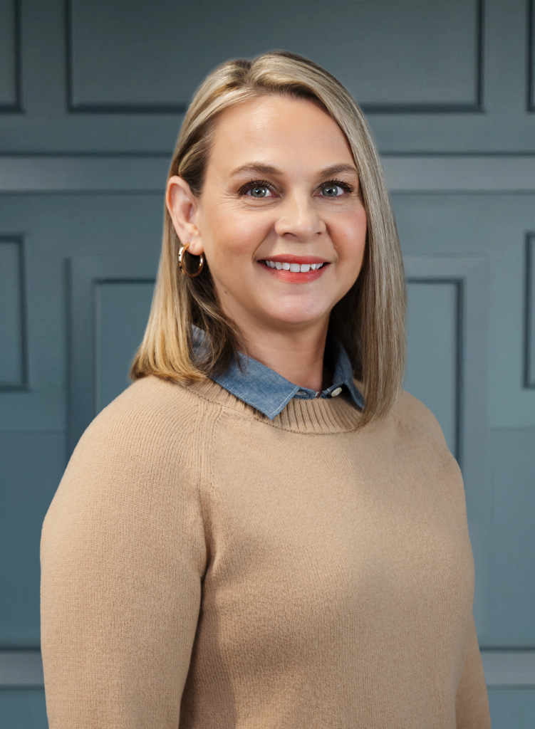 Smiling woman with shoulder-length blonde hair in a beige sweater against a paneled wall.
