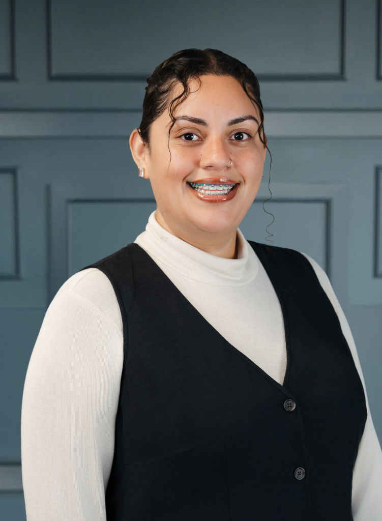 Smiling person with glasses and braces in a black vest over a cream turtleneck, posed against a blue paneled wall.