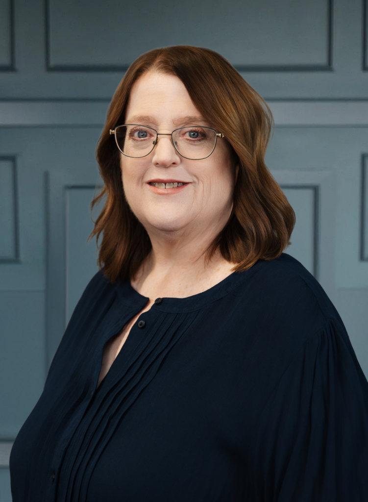 Smiling woman with glasses and brown hair wearing a blue blouse, posed in front of a blue paneled background.