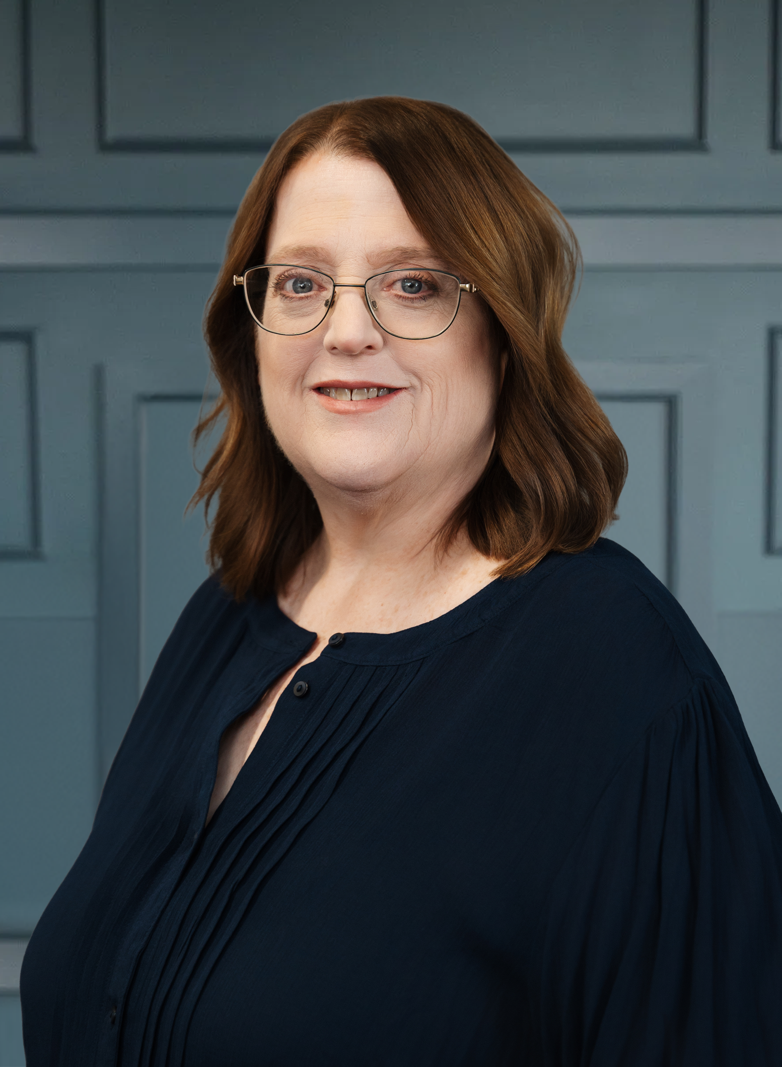 Smiling woman with glasses and brown hair wearing a blue blouse, posed in front of a blue paneled background.