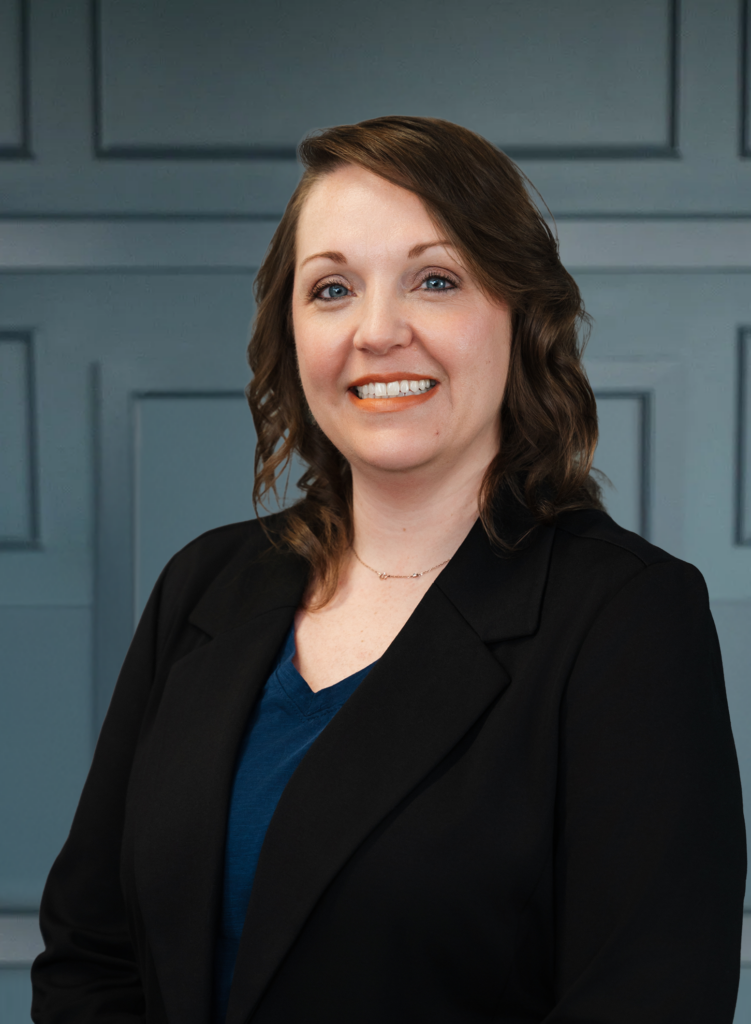 Smiling woman in a black blazer with a blue top, standing against a paneled wall.