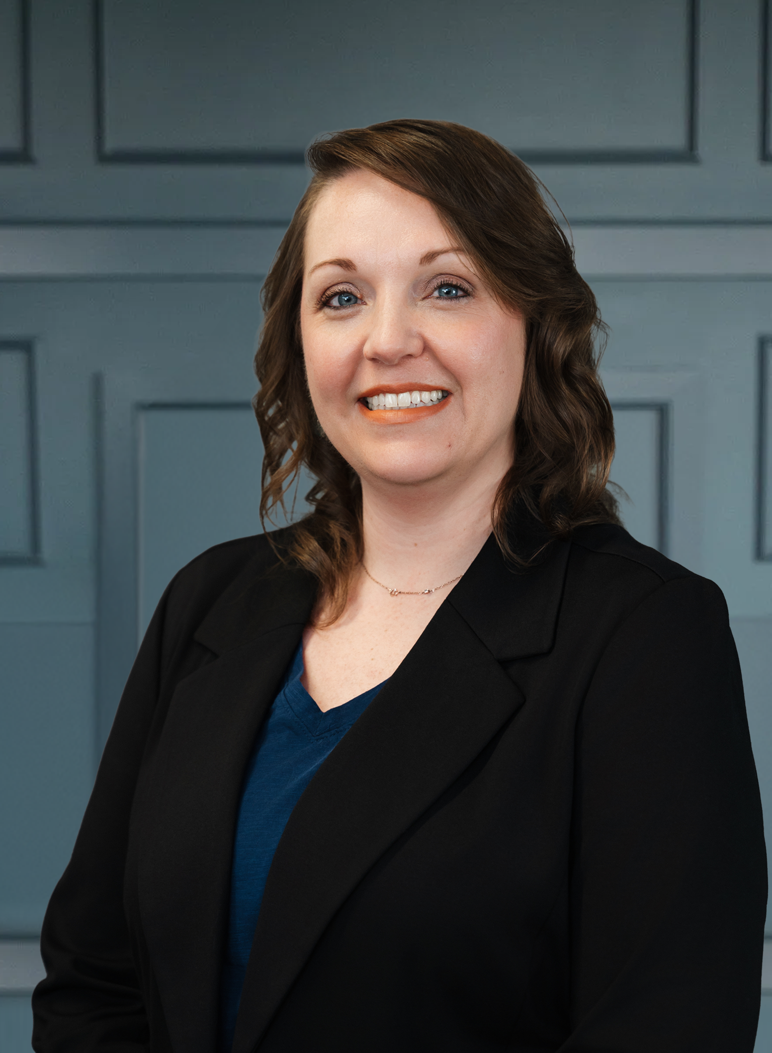 Smiling woman in a black blazer with a blue top, standing against a paneled wall.