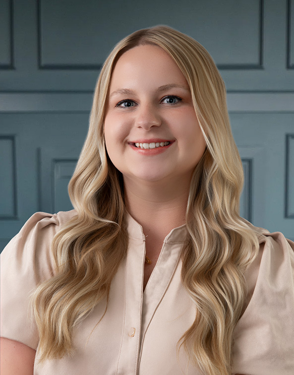 Smiling woman with long blonde hair wearing a beige blouse, standing against a blue paneled background.