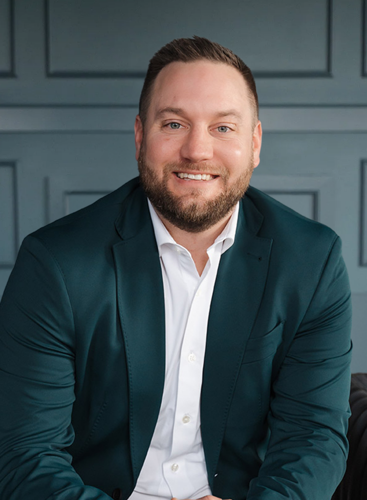 Smiling man in a dark blazer and white shirt, sitting against a paneled background.