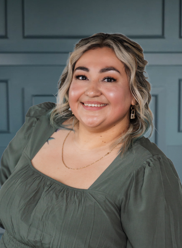 Smiling person with wavy blonde hair in a green top, posing against a paneled blue background.