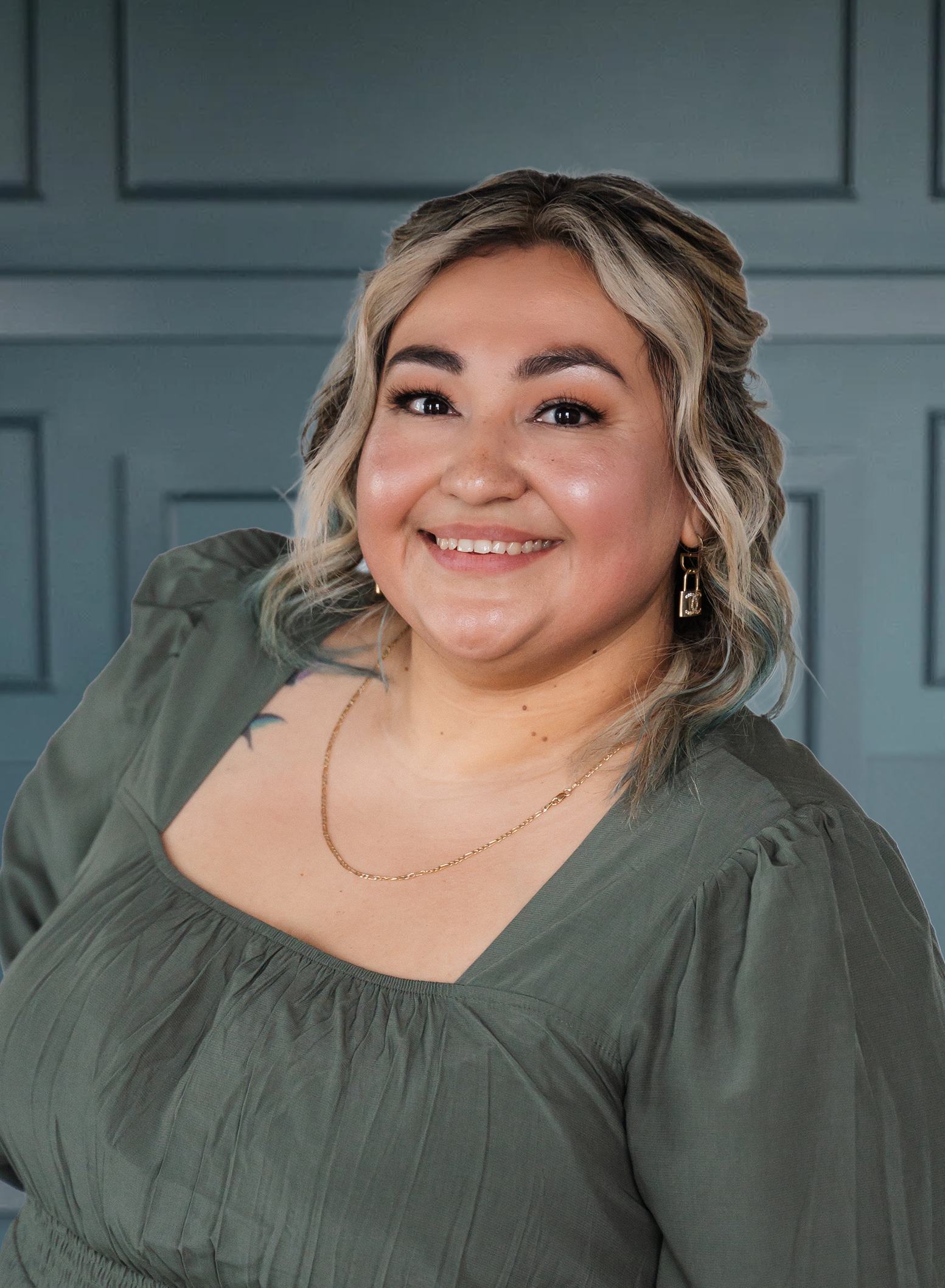 Smiling person with wavy blonde hair in a green top, posing against a paneled blue background.