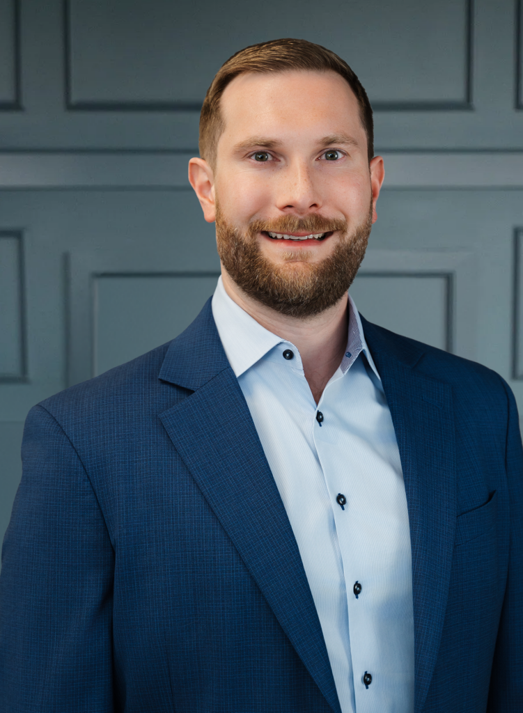 Smiling man in a blue suit and white shirt, standing against a paneled gray background.