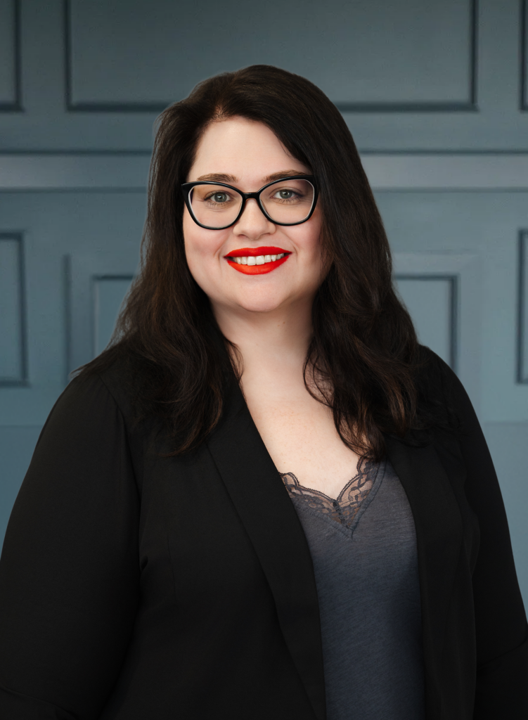 Woman with long dark hair, wearing black glasses and red lipstick, in a black blazer and lace top, smiling against gray wall.