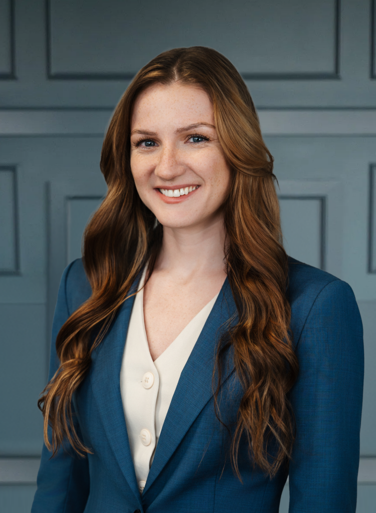 Smiling woman with long hair in a blue blazer standing against a paneled wall.