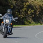Motorcyclist in black helmet and gear riding a classic cruiser on a curvy road bordered by trees.