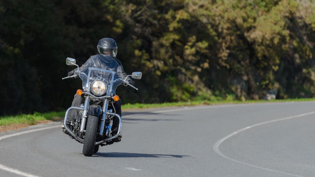 Motorcyclist in black helmet and gear riding a classic cruiser on a curvy road bordered by trees.