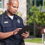 Police officer using a tablet outdoors near parked cars and buildings, focused on his task.