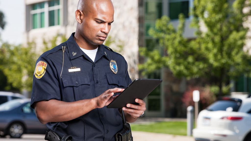Police officer using a tablet outdoors near parked cars and buildings, focused on his task.