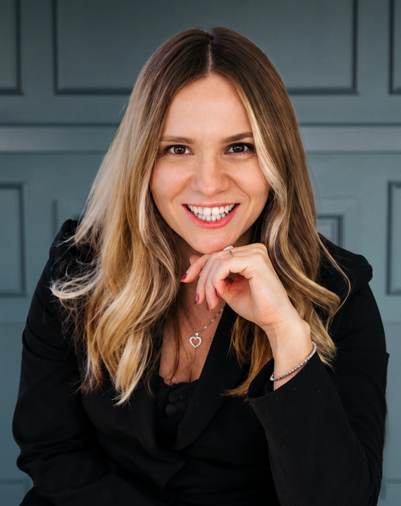 Smiling woman with long blonde hair in a black suit, posing in front of a blue paneled wall.