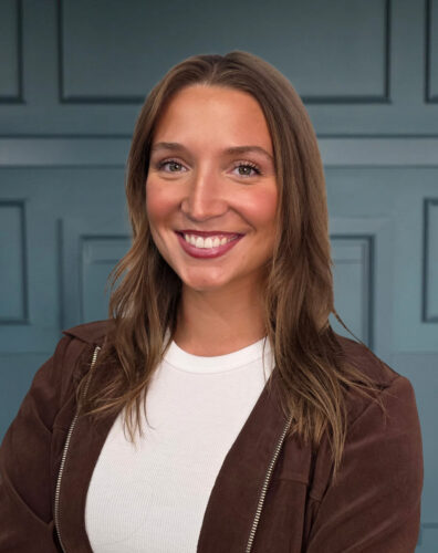 Smiling woman with long brown hair in a brown jacket, standing against a blue paneled wall.