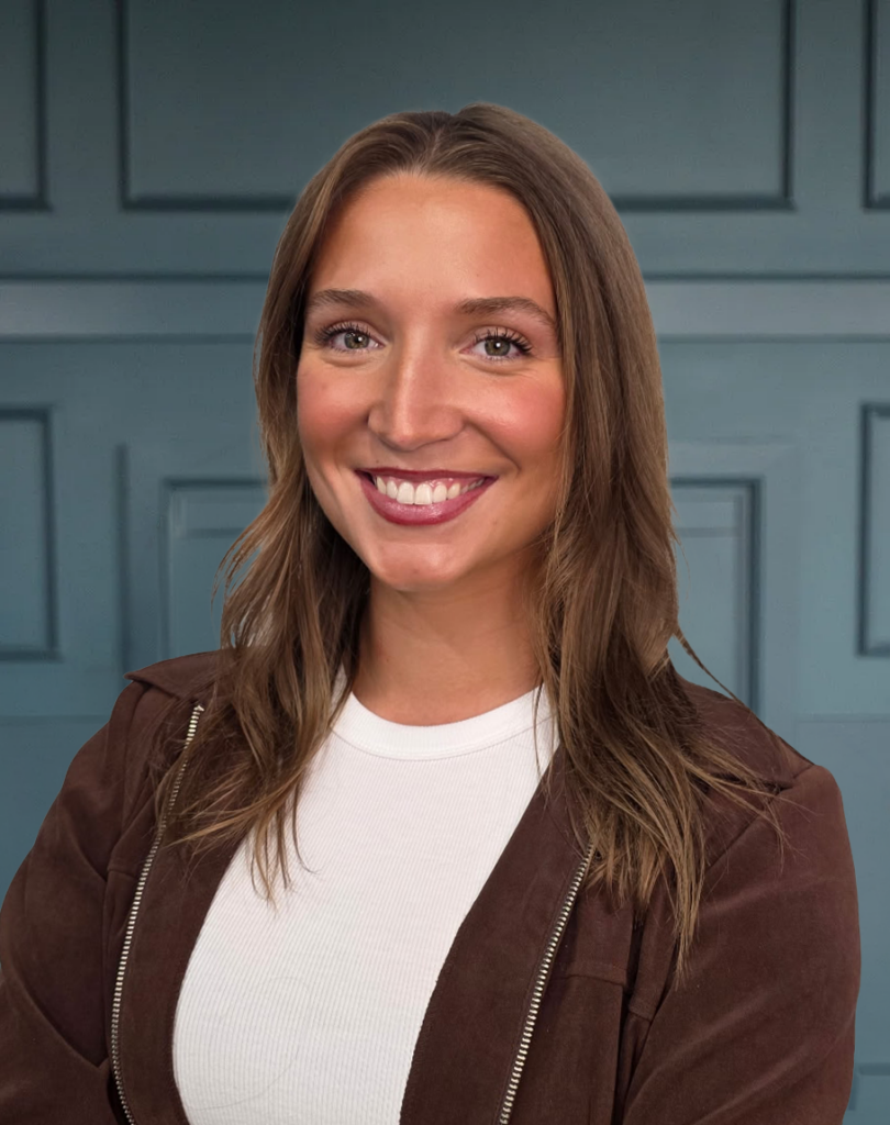 Smiling woman with long brown hair, dressed in a brown jacket and white top, standing against a blue paneled wall.